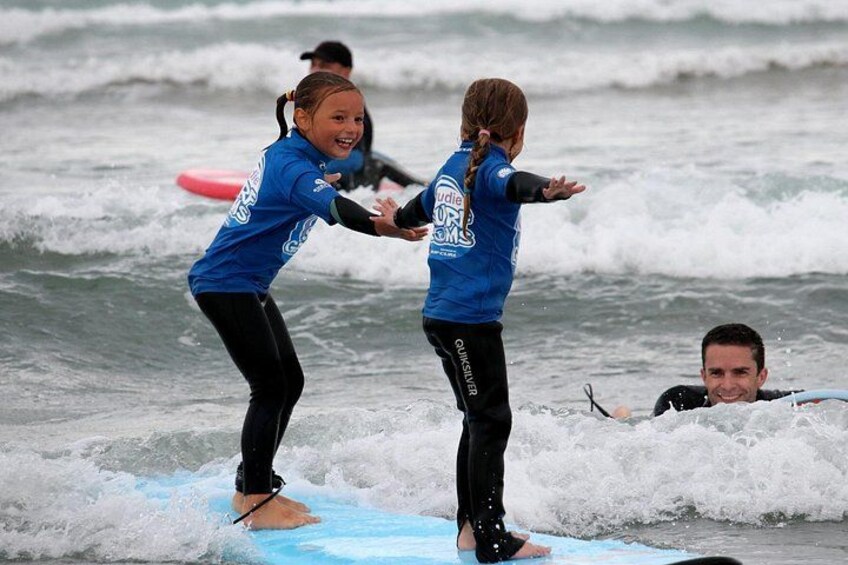 Twee kinderen in blauwe zwempakken staan op een surfplank in de oceaan, met golven eromheen. 2