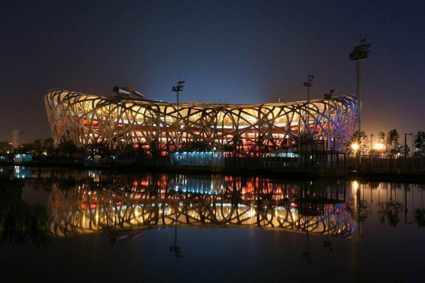 A large, illuminated stadium with a complex, circular design, reflecting on a calm body of water. 2