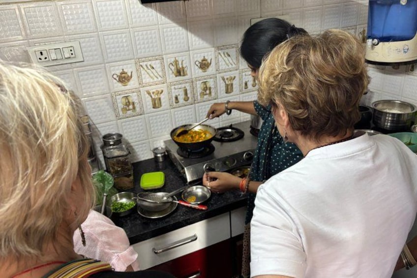 Una cocina con una persona cocinando en la estufa, otra persona de pie cerca y varios utensilios de cocina sobre la encimera. 4