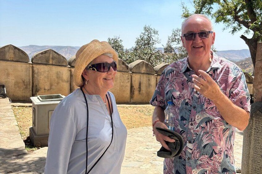 Two people standing outdoors with a stone wall and mountains in the background. 1
