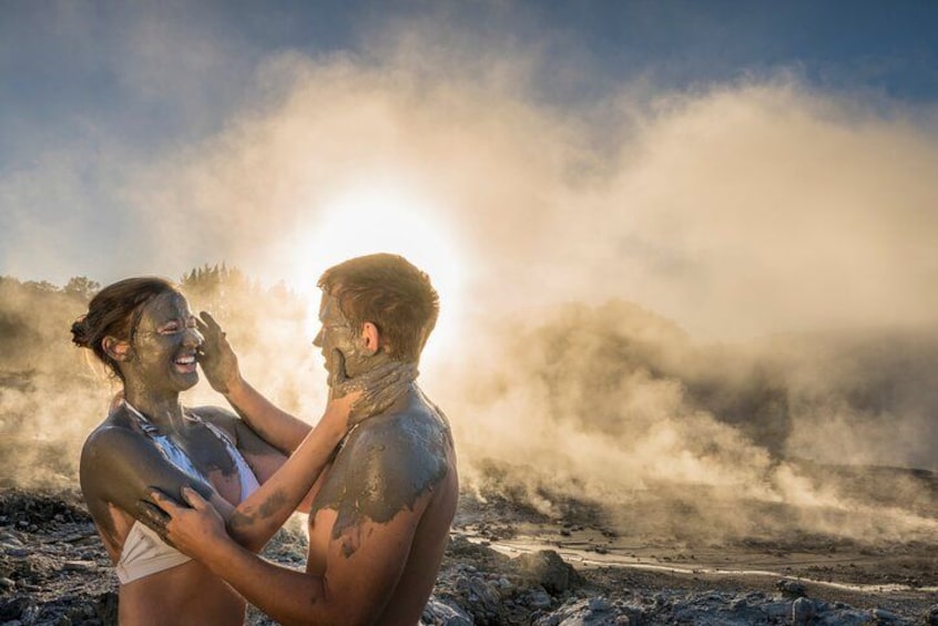 Two people covered in mud, standing on a beach with a misty background and the sun in the sky. 2