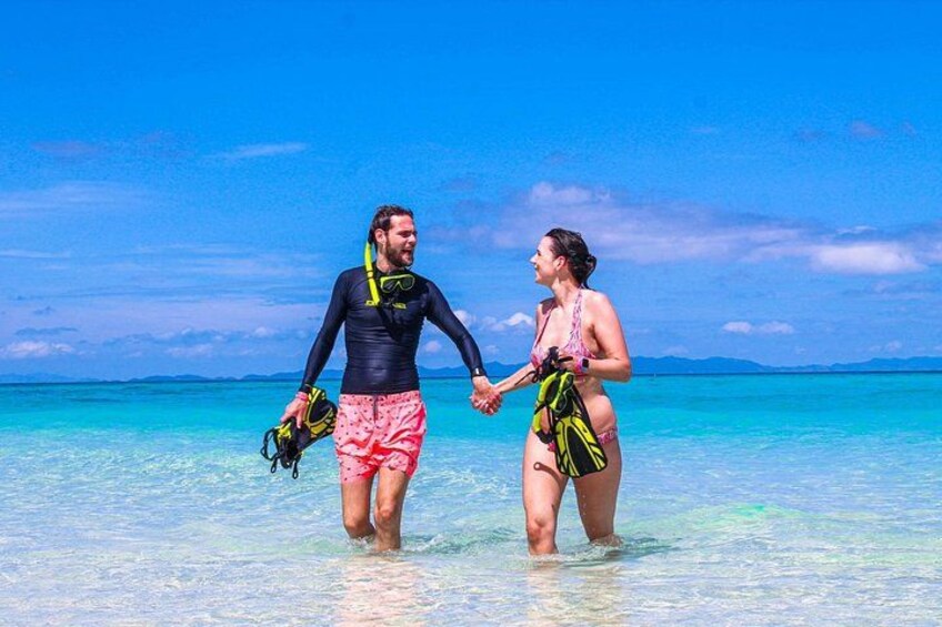Ein Paar spaziert Hand in Hand am Strand, mit klarem, blauem Wasser und einer entfernten Küstenlinie. 1