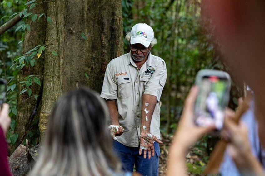 A man in a forested area, wearing a cap and a shirt with a logo, surrounded by trees and people taking photos. 4