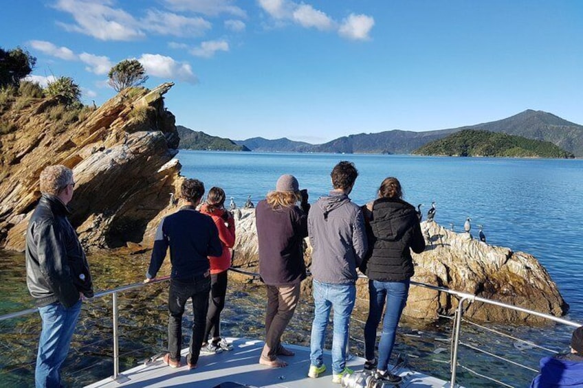 Un groupe de personnes se tient sur un bateau, près d’une côte rocheuse, avec un lac calme et des montagnes qui se dessinent à l’horizon. 4