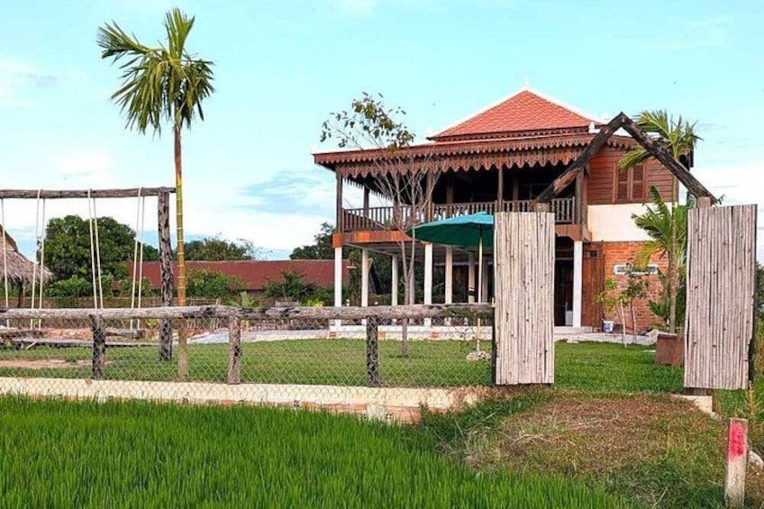 A wooden house with a red roof, surrounded by a fenced area with a palm tree and a grassy field. 5