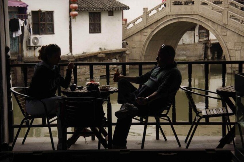 Two people sitting at a table on a balcony overlooking a river with a stone bridge in the background. 4