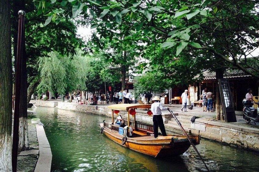 A narrow waterway with a wooden boat, people walking along the banks, and trees providing shade. 1
