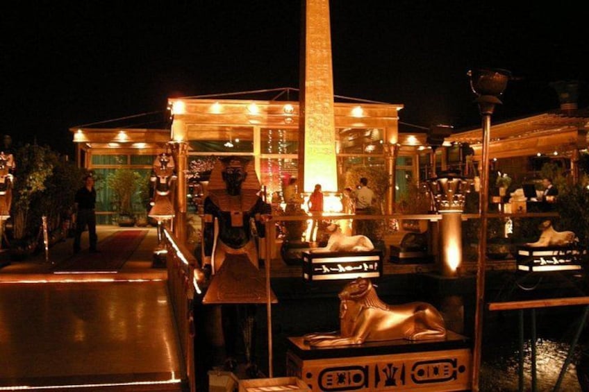 A nighttime outdoor market with illuminated stalls, a central fountain, and decorative statues. 1