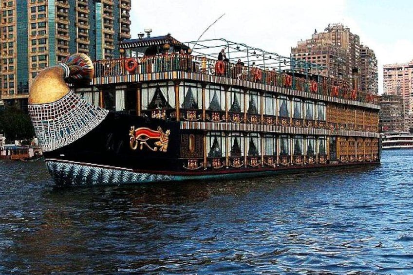 A large, ornate boat with a golden headpiece, decorated with hieroglyphs, floating on water with modern buildings in the background. 5