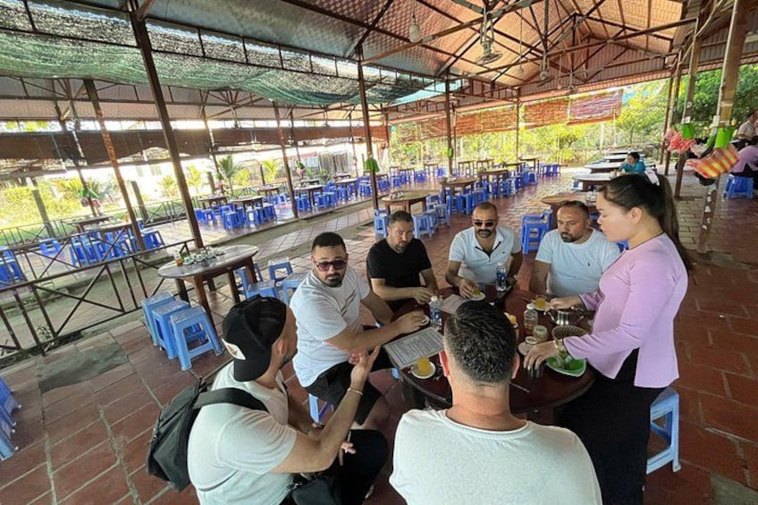 A group of people sitting around a table in an outdoor setting with blue plastic chairs and tables, under a covered area with a metal roof. 2