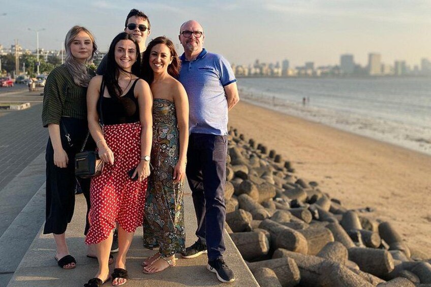 Un grupo de personas caminando por un sendero pavimentado junto a la playa, con un litoral rocoso y el perfil de una ciudad al fondo. 3