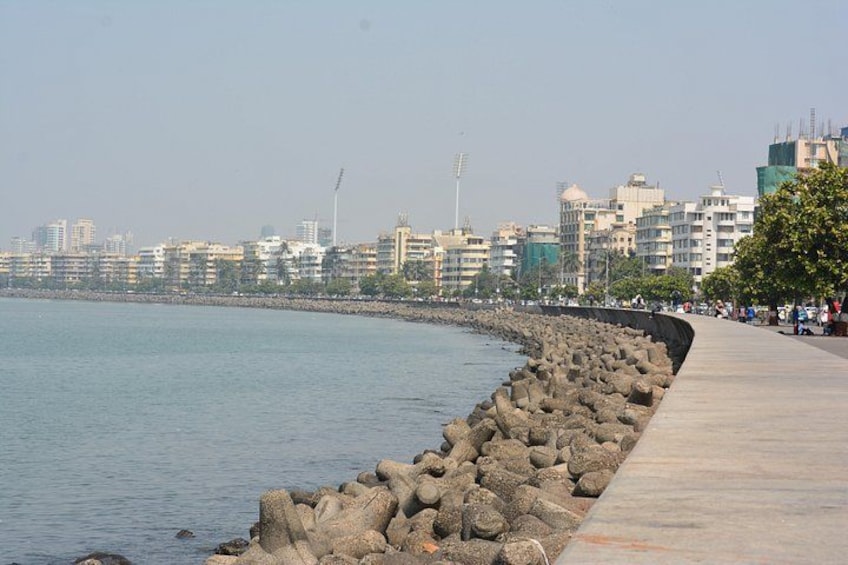 A waterfront promenade with a rocky breakwater, a city skyline in the background, and a clear sky. 1