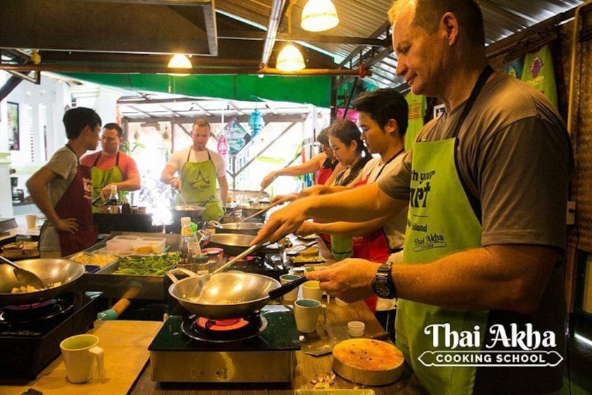 Un cours de cuisine est en cours, les participants portent des tabliers, des ustensiles de cuisine et un tablier vert sur lequel est inscrit le texte « Thai Akha Cooking School ». 4