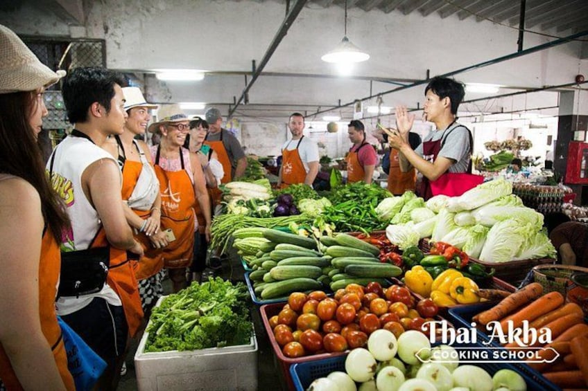 Un marché où l’on voit des gens portant des tabliers orange, une variété de légumes et une enseigne indiquant l’emplacement de l’école de cuisine thaïlandaise Akha. 1