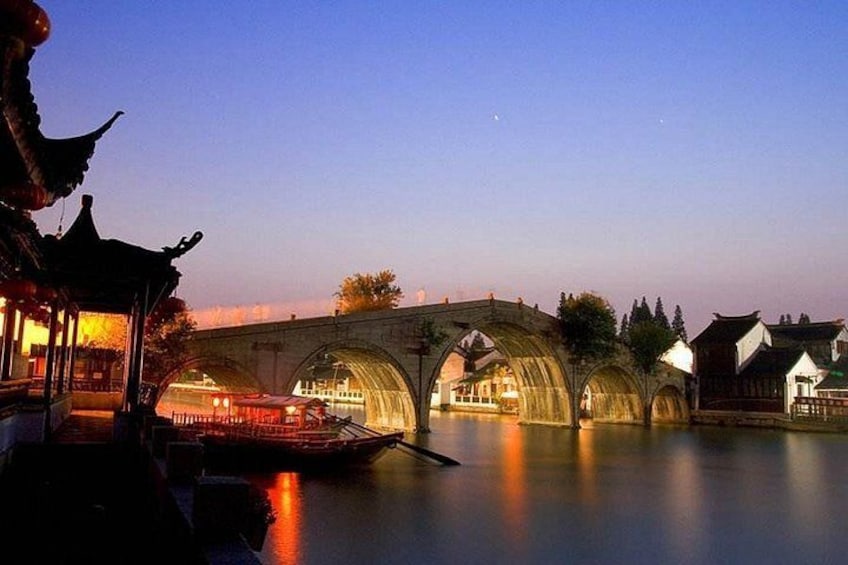 A traditional bridge over a calm river at dusk, with illuminated buildings and a boat. 4