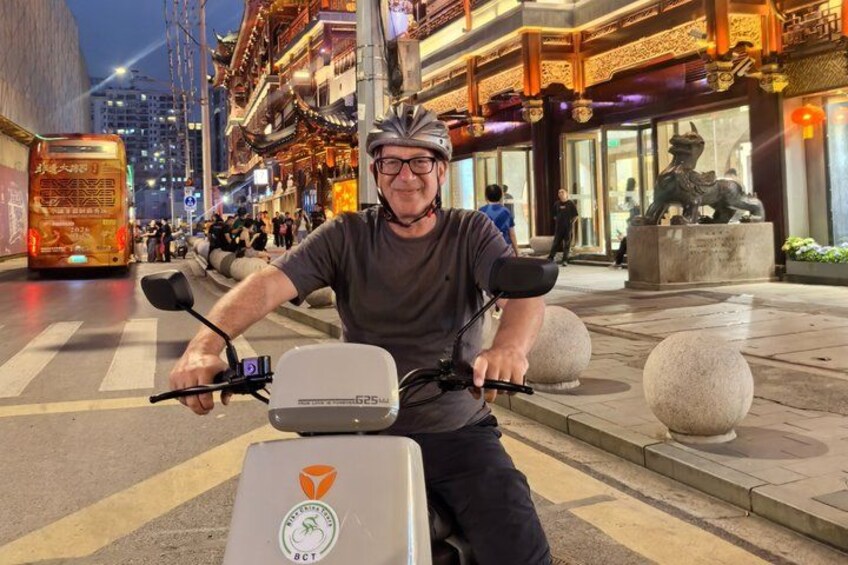 A person riding a scooter on a street with traditional architecture and a statue in the background. 5
