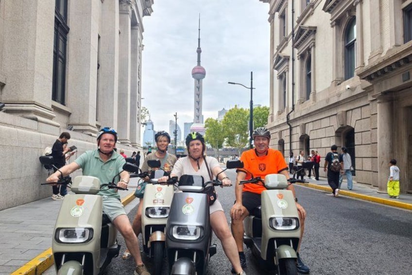 Four people on electric scooters on a city street with historic buildings and a tower in the background. 2