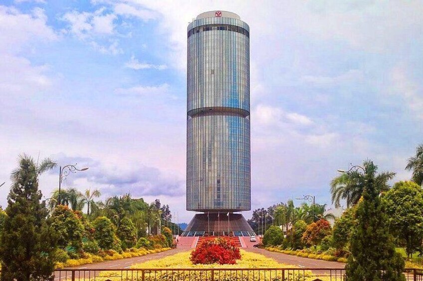 A tall, cylindrical skyscraper with a reflective glass facade, surrounded by well-maintained gardens and palm trees. 3