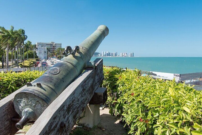 A cannon on display with a city skyline and ocean in the background. 1
