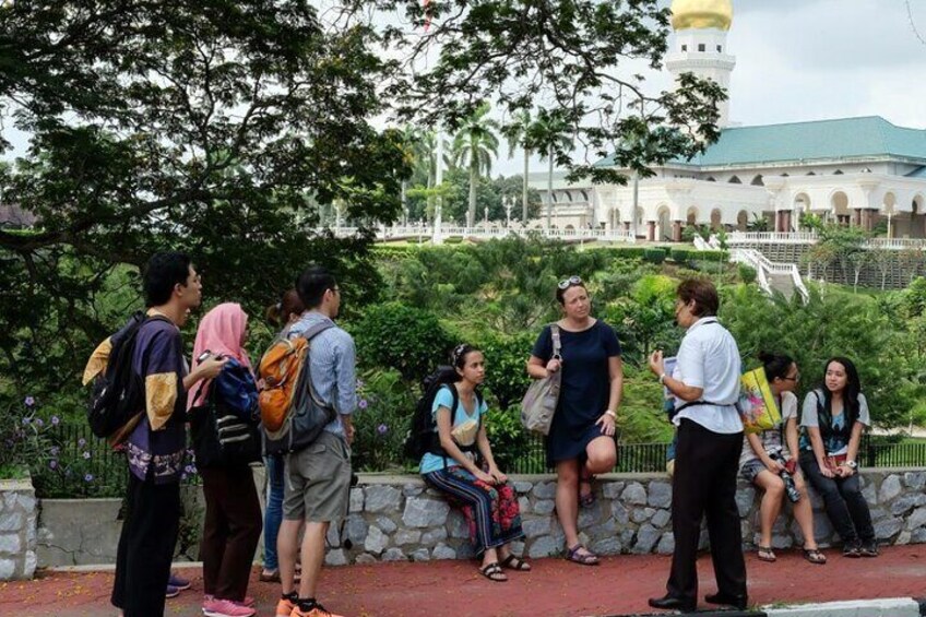 A group of people standing and sitting near a historical building with a green roof and white walls. 2