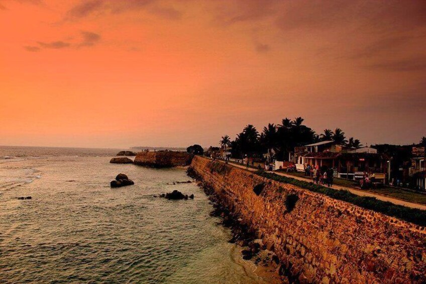 A coastal sunset with a stone wall, palm trees, and buildings along the shore. 3