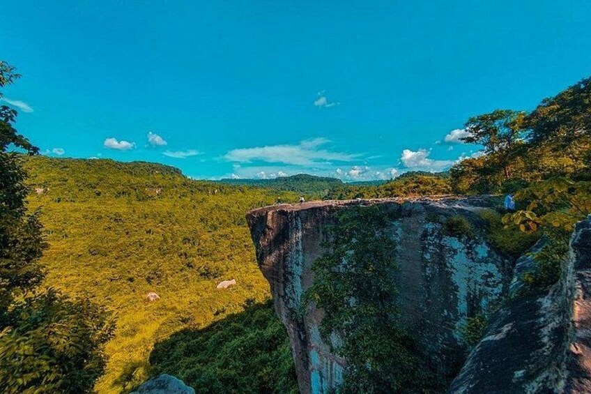 Un acantilado con una persona al borde, un exuberante valle verde abajo y un cielo azul despejado. 2