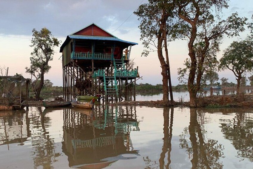 A house on stilts by a calm body of water, surrounded by trees. 2