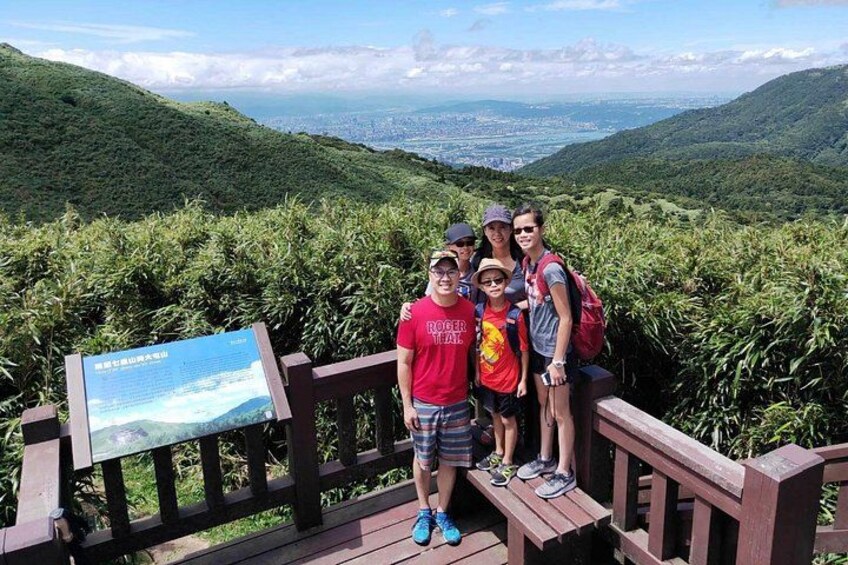 A group of people standing on a wooden platform with a scenic view of green hills and a distant cityscape. 3