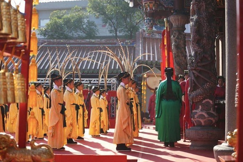 Een groep mensen in traditionele kleding staat in een rij, met een grote, versierde structuur op de achtergrond, en een persoon in het groen in de voorgrond. 5