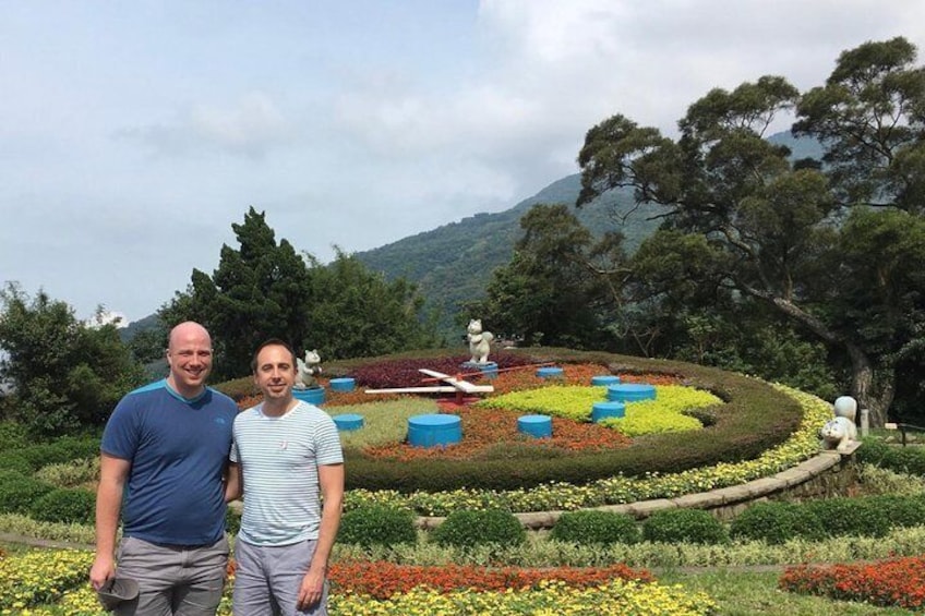 Two men standing in a garden with colorful flower beds and statues. 4