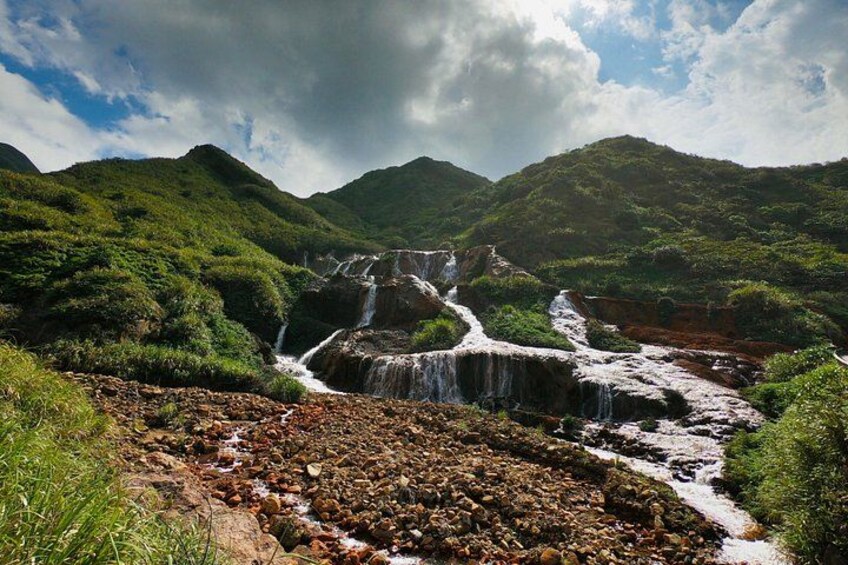 Una cascata che si getta su rocce in una rigogliosa valle verde, con colline sullo sfondo. 2