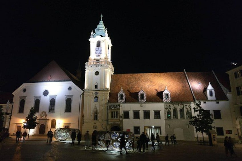 A historic building with a clock tower, illuminated windows, and a courtyard with people. 3