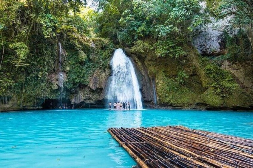 A clear blue pool with a wooden dock, a waterfall, and lush greenery. 2