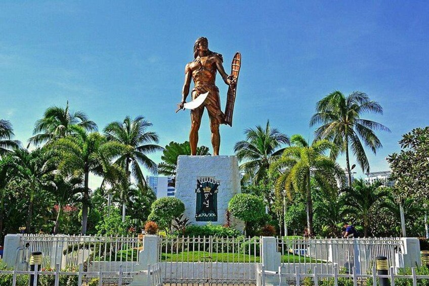 Eine Statue einer muskulösen Figur, die Schild und Speer hält, umgeben von Palmen und einem Zaun. 5