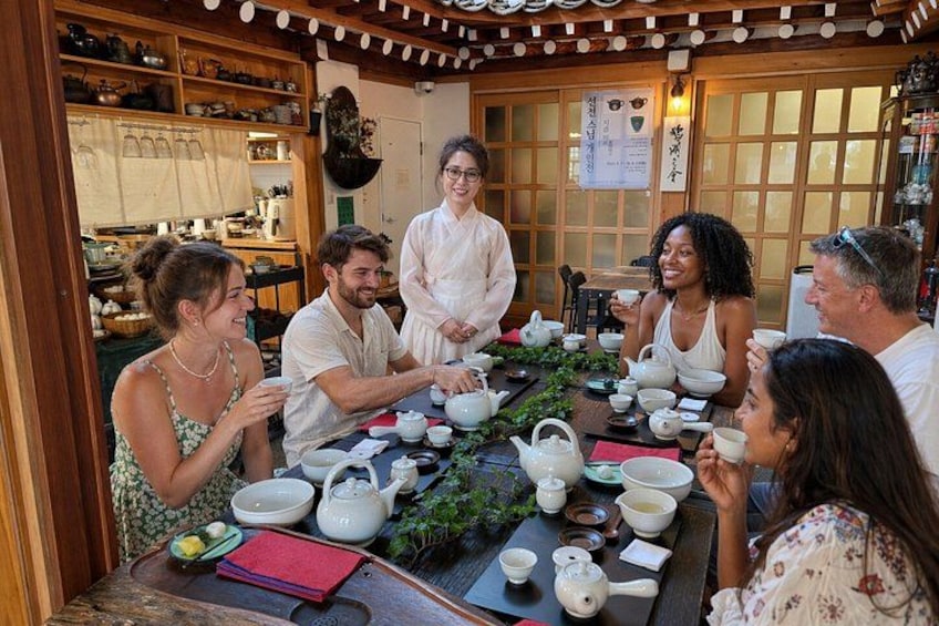 A group of people gathered around a table with teapots, cups, and plates, in a room with wooden furniture and a traditional Japanese aesthetic. 2