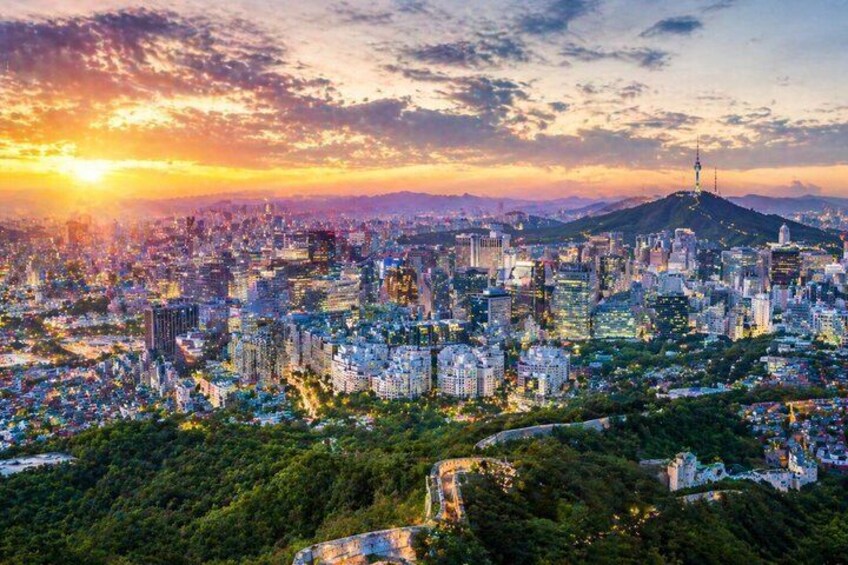 Un paisaje urbano al atardecer con una torre destacada en una colina, edificios urbanos densos y un área boscosa en primer plano. 5