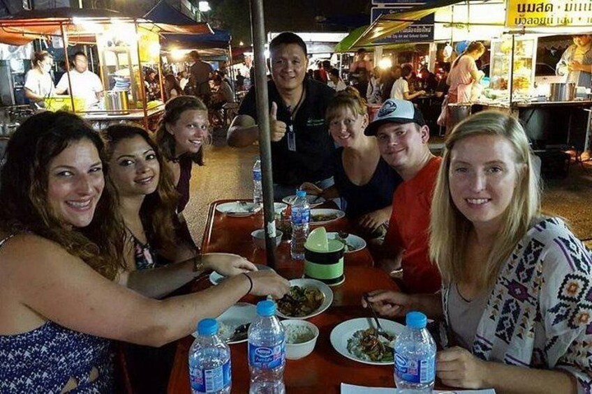 A group of people dining at an outdoor market with food stalls in the background. 5