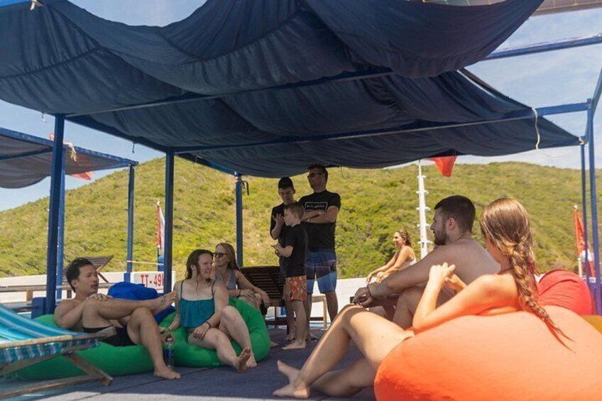Personas relajándose en sillas de playa y pufs bajo una zona cubierta, con montañas al fondo. 2