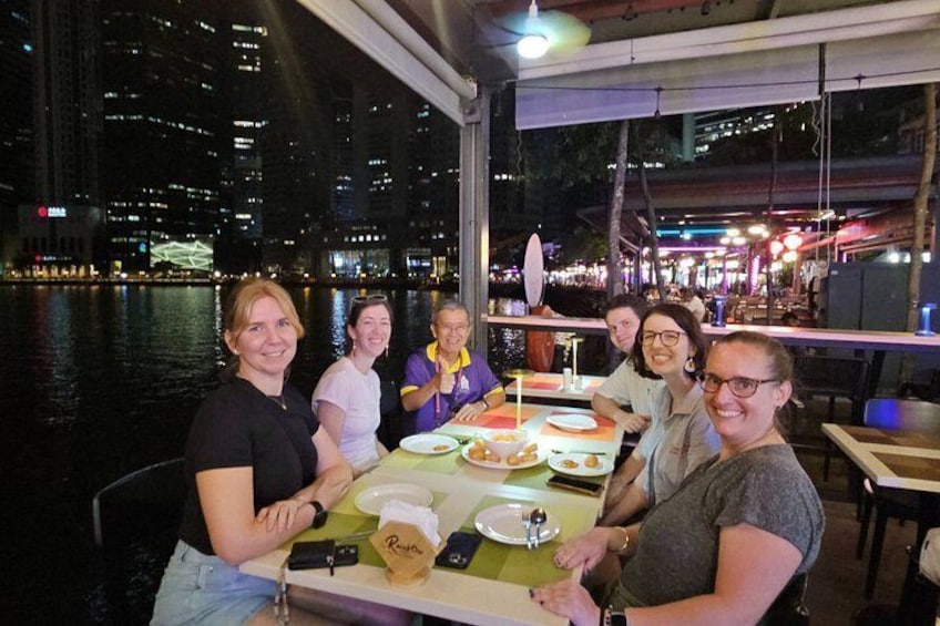 Un grupo de personas cenando en un restaurante frente al mar, con un paisaje urbano de fondo. 3