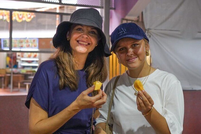 Two people holding food items, with a food stall and a purple wall in the background. 2