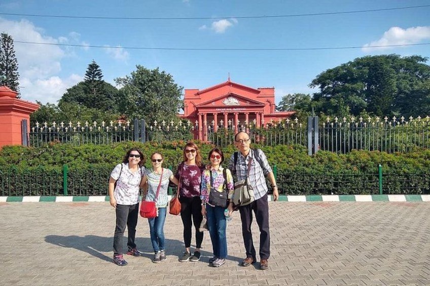 A group of people standing in front of a red building with columns and a fence with greenery behind. 5