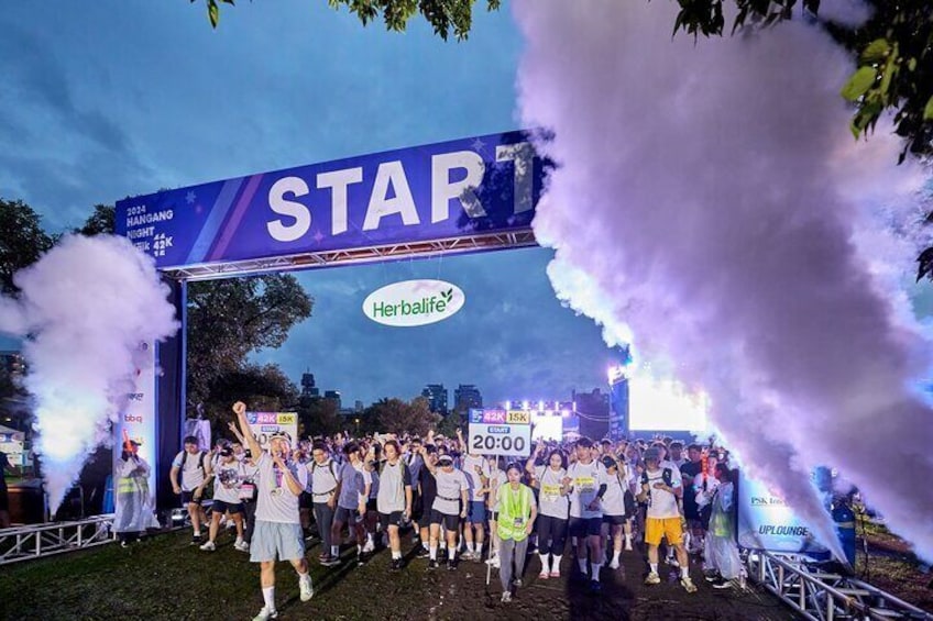 A group of people at a starting line with a 'START' arch and 'Herbalife' branding. 1