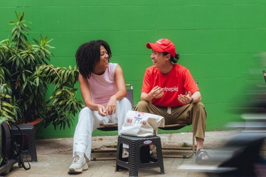 Two people sitting on a bench, one wearing a red shirt and the other in a pink top, with a green wall and potted plants in the background. 3