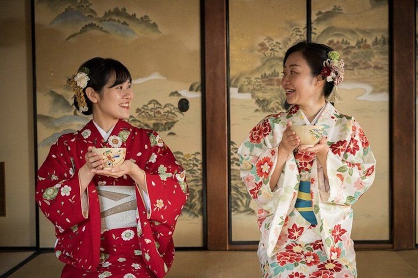 Zwei Personen in traditionellen japanischen Kimonos mit Blumenmustern, vor einer Landschaftsdarstellung stehend. 4