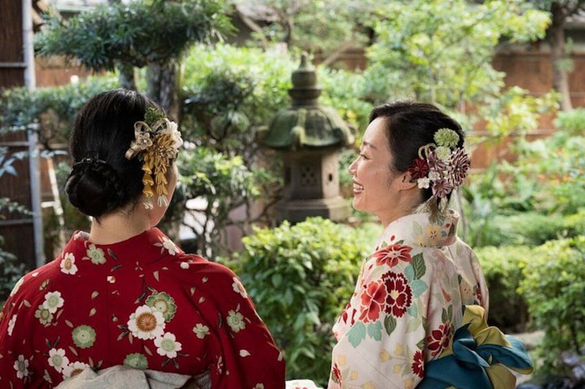 Two individuals in traditional attire with floral patterns, standing in a garden with greenery and a stone lantern in the background. 4