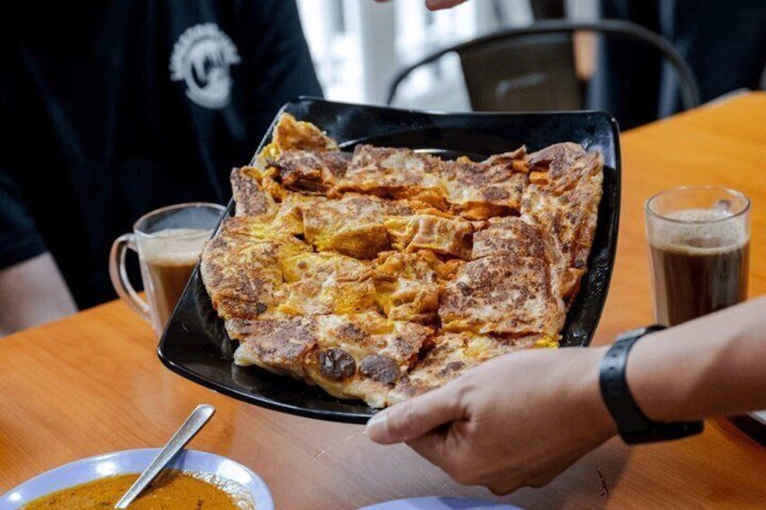 A person serving a large, rectangular, layered dish on a black plate, with a bowl of sauce and a cup of coffee on the table. 4