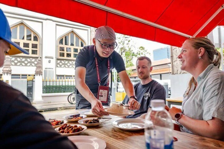 A group of people dining at an outdoor table with a red canopy, under a building with arched windows. 1