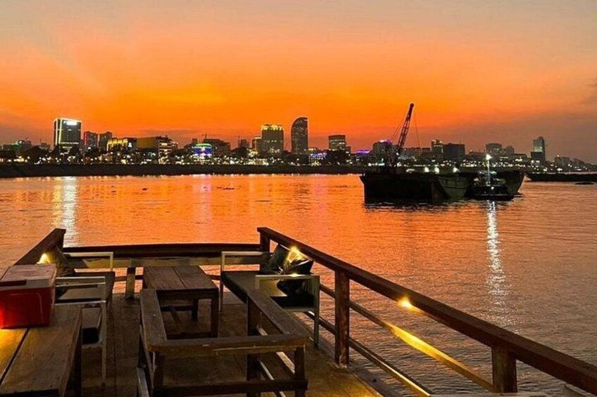 Une vue sur le coucher de soleil depuis une jetée, avec des bancs en bois et une rambarde, d’où l’on aperçoit la silhouette d’une ville et un grand navire sur l’eau. 5