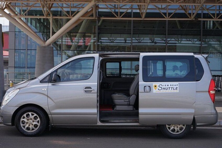 En sølvfarvet shuttlebus med teksten 'Shuttle' og 'IEM REAP' på siden, parkeret foran en moderne bygning med glasfacade. 2