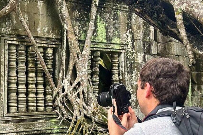 Eine Person fotografiert eine Steinstruktur mit aufwendigen Schnitzereien und überwuchernden Baumwurzeln. 3